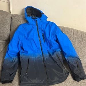 Blå och svart jacka från Northville - Säljer en cool blå och svart jacka från Northville. Jackan har en gradvis färgövergång från blått till svart och är utrustad med dragkedja och flera fickor. Perfekt för kyligare väder med sin vindtäta och vattentäta design. Huvan är justerbar och insidan är fodrad för extra värme.