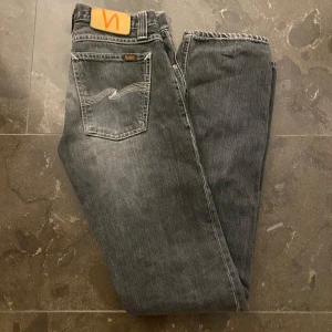 Grå jeans från Nudie - Snygga grå jeans från Nudie med klassisk femficksdesign och kontrasterande sömmar på bakfickorna. Jeansen har wn tvär snygg tvätt. Tyvärr va de för små för mig och jag är 180cm. Skulle säga att de passar folk runt 170cm och folk som har lite mindre midja!👌Bara o höra av sig vid frågor!