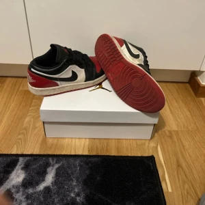 Nike Air Jordans i svart, röd och vit - Snygga Nike Air Jordans i en klassisk färgkombination av svart, röd och vit. Skorna har en låg profil med snörning och den ikoniska swoosh-loggan på sidan. Perfekta för en stilren look.