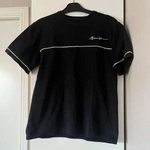 Svart t-shirt med vit broderad text - Snygg svart t-shirt med vit broderad text på bröstet och gröna linjer över bröstet och ärmarna. Tjockare material. 