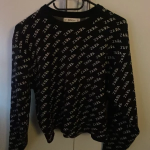 Svart stickad tröja från Zara - Säljer en svart stickad tröja från Zara med ett vitt logotypmönster över hela plagget. Tröjan har långa ärmar och en rund halsringning. Perfekt för en trendig och bekväm look. 🖤