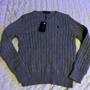 Grå stickad tröja från Ralph Lauren - Säljer en stilren grå stickad tröja från Ralph Lauren med klassiskt kabelstickat mönster. Tröjan har en v-ringad hals och långa ärmar, perfekt för kyligare dagar. Den ikoniska loggan är broderad på bröstet. Ett måste i garderoben för en tidlös look.