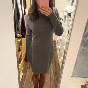 Grå ribbad klänning med knappar - Snygg grå ribbad klänning med guldknappar längs sidan och axeln. Den har en tight passform och långa ärmar, perfekt för kyligare dagar. Klänningen är stilren och enkel att matcha med olika accessoarer. 