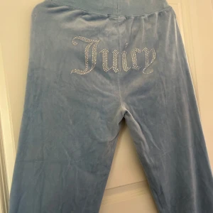 Ljusblå velourbyxor från Juicy Couture - Snygga ljusblå velourbyxor från Juicy Couture med glittrande stenar som bildar texten 'Juicy' på baksidan. Byxorna har en bekväm passform och snörning i midjan för justerbarhet. Perfekta för en avslappnad stil med en touch av glamour. Byxorna är sparsamt använda samt i fint skick. Nypris för dessa är 1100kr, men jag säljer för 500kr. Vid frågor eller funderingar hör av er, jag svarar så fort jag kan!🩷