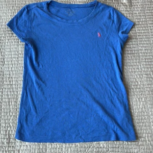 Blå t-shirt från Ralph Lauren - Snygg blå t-shirt från Ralph Lauren med en liten rosa logga på bröstet. Perfekt för en avslappnad stil. T-shirten har en rund hals och korta ärmar.