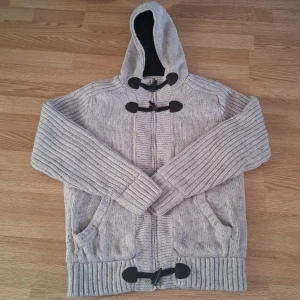 Grå stickad jacka med huva från Angelo Litrico - Säljer en grå stickad jacka från Angelo Litrico med huva och svarta knappar. Jackan har en mysig, mörk insida och ribbade muddar. Perfekt för kyliga dagar!