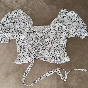 Vit prickig topp från Shein - Säljer en söt vit topp med svarta prickar från Shein. Toppen har korta puffärmar och en smockad design med knytband i midjan. Perfekt för en trendig look!