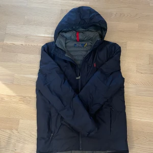 Mörkblå dunjacka från Polo Ralph Lauren - Säljer en snygg mörkblå dunjacka från Polo Ralph Lauren. Jackan har en huva och dragkedja framtill. Den är perfekt för kyligare dagar och har en diskret logga på bröstet. Passar bra för både höst och vinter.