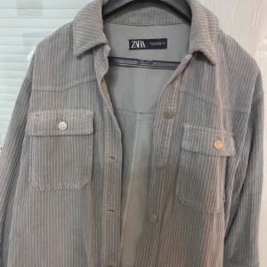Grå overshirt från Zara - Säljer en stilren grå overshirt från Zara med diskreta ränder. Den har långa ärmar och två bröstfickor med knappar. Perfekt för lager-på-lager-stil. Passar bra till både casual och mer uppklädda tillfällen.