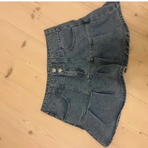 Blå jeanskjol med knappar - Snygg blå jeanskjol med två dekorativa knappar framtill. Kjolen har en kort modell och är perfekt för en avslappnad stil. Den har en klassisk jeanslook med fickor och sömmar som ger en cool touch.