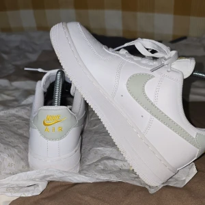 Vita Nike Air Force med gulddetaljer - Snygga vita Nike Air Force med gulddetaljer och en ljusgrå swoosh. Skorna har klassisk snörning och perforeringar på tån för ventilation. Perfekta för en stilren look.