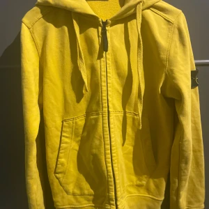 Gul huvtröja från Stone Island - Säljer en snygg gul huvtröja från Stone Island med dragkedja och huva. Tröjan har en klassisk look med ribbade muddar och en logotyp på ärmen. Perfekt för en casual stil.