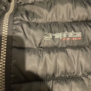 Svart dunväst från Graphite Sportswear - Snygg svart dunväst från Graphite Sportswear med dragkedja framtill. Perfekt för lager-på-lager under kyliga dagar. Västen har en stilren design med diskret logga på bröstet.