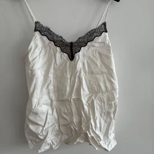 Vit linne med spetsdetalj - Säljer en vit linne med smala axelband och svart spetsdetalj vid halsringningen. Perfekt för en stilren och elegant look. Passar bra till både jeans och kjol.