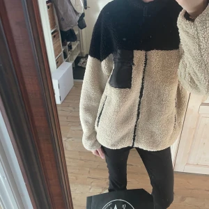 Beige teddyjacka från Gina Tricot - Mysig beige teddyjacka från Gina Tricot med svarta detaljer. Jackan har en hög krage och dragkedja framtill. Den har en praktisk ficka med dragkedja på bröstet och är perfekt för kyligare dagar.