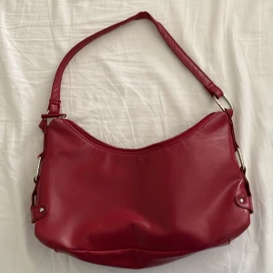 Röd axelväska i syntet - Snygg röd axelväska i syntetmaterial med en praktisk dragkedja upptill. Väskan har en enkel design med metallspännen på sidorna som ger en stilren look. Perfekt för att bära med sig det viktigaste i vardagen.