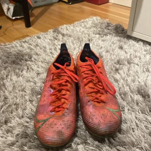 Röda Nike Mercurial fotbollsskor Elite 2021 - Säljer ett par röda Nike Mercurial fotbollsskor med snörning och blå detaljer. Skorna har en slitstark sula med dobbar för bra grepp på planen. Perfekta för fotbollsspelare som vill ha både stil och funktion. Lite sönder i hälen