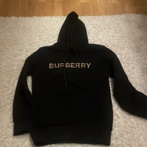 Svart hoodie från Burberry - Säljer en stilren svart hoodie från Burberry med deras ikoniska logga i fram. Tröjan har en klassisk känguruficka och justerbar huva med dragsko. Perfekt för en avslappnad och trendig look.