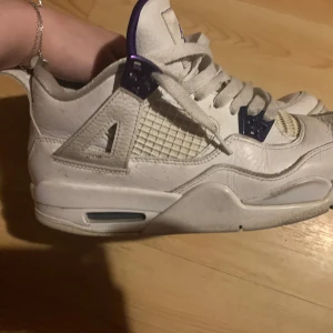 Vita och lila Nike Air Jordans - Snygga vita Nike Air Jordan 4s i modellen metallic purple. Skorna har en klassisk design med snörning och en högre siluett. Perfekta för dig som vill ha en stilren och sportig look.