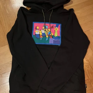 Svart dress med Simpsons-tryck från Billabong - Säljer en svart hoodie samt byxor från Billabong med ett färgglatt Simpsons-tryck på framsidan. Hoodien har en justerbar huva med mönstrade snören och en stor ficka framtill. Byxorna har Simpson-tryck på vardera ben. Perfekt för alla Simpsons-fans! 🖤 Byxorna i storlek 16 (174) och tröjan i S.