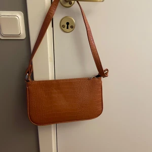 Brun axelväska med krokodilmönster - Snygg brun axelväska i syntetmaterial med krokodilmönster. Väskan har en praktisk dragkedja och en justerbar axelrem. Perfekt för att ge din outfit en trendig touch.