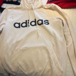 Vit hoodie från Adidas - Säljer en vit hoodie från Adidas med svart logga på bröstet. Den har en klassisk känguruficka och justerbar huva med dragsko. Perfekt för en avslappnad stil.