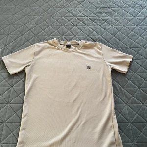 Beige t-shirt från Burbery - Säljer en stilren beige t-shirt från Burbery. Den har en liten broderad logga på bröstet och är tillverkad i ett mjukt material. Perfekt för en avslappnad look.