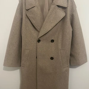 Beige kappa från Zara - Säljer en stilren beige kappa från Zara med dubbelknäppning och stora slag. Kappan har en klassisk design med två fickor framtill och är perfekt för kyligare dagar. Är lite nopprig men inte något som synd (kan tas bort)