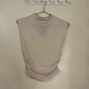 Beige ärmlös topp - Säljer en stilren beige ärmlös topp med hög hals. Toppen har en elegant drapering på sidan som ger en snygg detalj. Perfekt för att matcha med olika outfits. Den är lite genomskinlig. Lappen är fortfarande kvar på, den är helt ny.