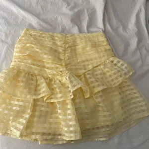 Gul rutig kjol från Gina Tricot - Säljer en söt gul rutig kjol från Gina Tricot med volangdetaljer. Kjolen har en dragkedja på sidan och är perfekt för sommarens varma dagar. Den har en kort längd och ett lekfullt utseende med sina volanger.