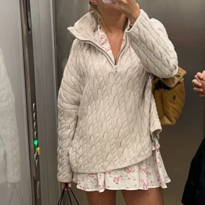 Tröja - Mysig beige stickad tröja med dragkedja och kabelstickat mönster. Perfekt för kyliga dagar med sin avslappnade passform och långa ärmar. Tröjan har en hög krage som kan bäras uppvikt eller nedvikt.