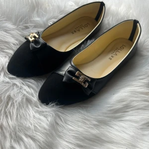 Svarta ballerinaskor från Lolla - Eleganta svarta ballerinaskor från Lolla med dekorativ rosett och guldfärgad detalj framtill. Skorna har en spetsig tå och är tillverkade i syntetmaterial, vilket ger en stilren look.