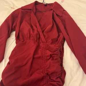 Röd omlottklänning från Shein - Jag har en snygg röd omlottklänning från Shein med långa ärmar och en smickrande v-ringning. Klänningen har en dragkedja på sidan och rynkade detaljer som ger en fin passform. Jag har aldrig använt den, men tyvärr är dragkedjan trasig. Om någon kan fixa den går den att använda, för den är verkligen jättefin – det är bara det här problemet med dragkedjan.