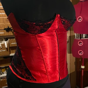 Röd korsett med spetsdetaljer - röd korsett med svarta spetsdetaljer och snörning bak. obs kuporna går ej hela vägen över bysten. stl 2XL men kan justeras med snörningen. har borttagbara strumpbandhållare!