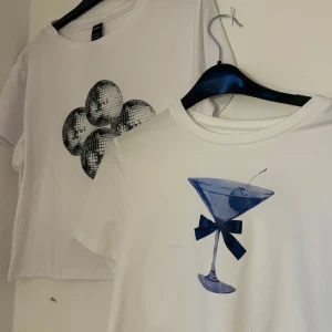 Två vita t-shirts med tryck - Säljer två vita t-shirts med tryck. Den ena har ett stort martiniglas med en blå rosett, och den andra har ett tryck av en discokula i form av en blomma. Perfekta för en avslappnad stil med en twist. 30kr för båda två. Storlek XL men är så pass små att de passar xs/s