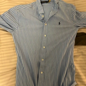 Blå och vit randig skjorta från Ralph Lauren - Snygg blå och vit randig skjorta från Ralph Lauren med klassisk krage och knappar framtill. Skjortan har en broderad logotyp på bröstet och är perfekt för en stilren look.