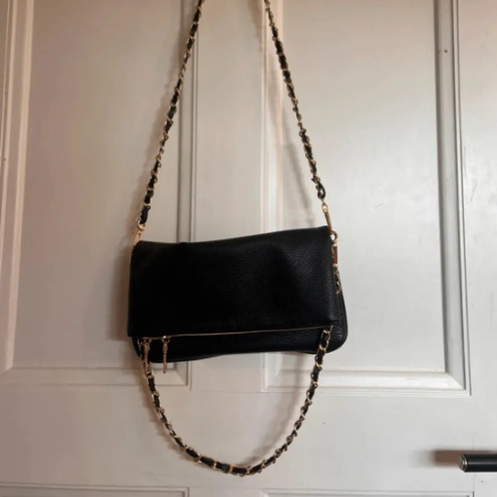 Snygg svart axelväska med guldfärgade kedjedetaljer på axelremmen. Väskan har en dragkedja framtill och är perfekt för att bära dina nödvändigheter med stil. Den har en elegant och tidlös design som passar till många tillfällen.. Laukut & Käsilaukut.
