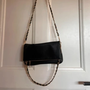 Svart axelväska med kedjedetaljer - Snygg svart axelväska med guldfärgade kedjedetaljer på axelremmen. Väskan har en dragkedja framtill och är perfekt för att bära dina nödvändigheter med stil. Den har en elegant och tidlös design som passar till många tillfällen.