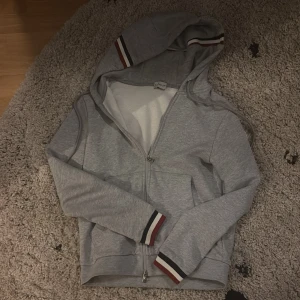 Grå huvtröja från Moncler - Säljer en stilren grå huvtröja från Moncler med dragkedja och randiga detaljer i svart, vit och röd på ärmslut och huva. Perfekt för en avslappnad look. Tröjan har långa ärmar och en bekväm passform. Koftan är i storlek S men passar Xs (små i storlekar)