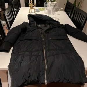 Svart dunjacka från Everest - Säljer en lång svart dunkändla jacka från Everest med huva och dragkedja. Reflexslitsar Jackan har långa ärmar och praktiska fickor framtill. Perfekt för kyliga dagar.