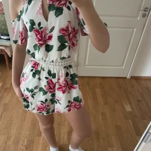 Vit blommig one piece med öppna axlar - Säljer en söt vit one piece med stora rosa blommor och gröna blad. One piecen har öppna axlar och en fin spetsdetalj i midjan. Perfekt för sommarens varma dagar!