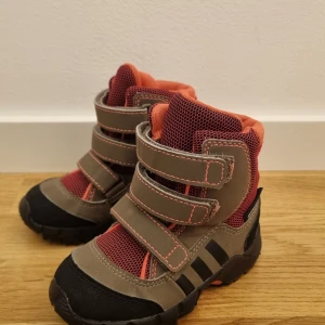 Barnboots från Adidas med kardborreband - Säljer ett par robusta barnboots från Adidas i grått och orange. Skorna har praktiska kardborreband för enkel på- och avtagning samt en slitstark sula för bra grepp. Perfekta för utomhusaktiviteter under höst och vinter.
