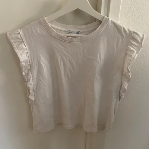 Vit topp med volangdetaljer - Säljer en söt vit topp med volangdetaljer vid ärmarna. Toppen är kortärmad och har en rund halsringning. Perfekt för en avslappnad stil.