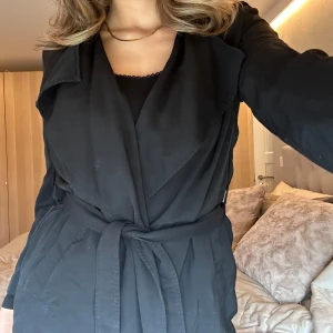 Svart kappa med knytband - Elegant svart kappa/kofta med knytband i midjan. Perfekt för en stilren look. Långärmad design som passar bra till både vardag och fest. Tunn