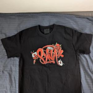 Syna World - Red Devil Tshirt (Ny) - Cool svart t-shirt med ett stort rött och vitt tryck på framsidan och baksidan. Designen har en djärv och edgy stil med text och grafiska element. Perfekt för en avslappnad och trendig look.