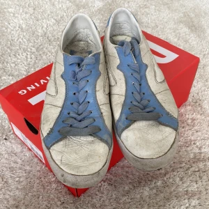 Vita och blå sneakers från Diesel - Säljer ett par vita sneakers från Diesel med blå detaljer och snörning. Skorna har en unik design med blå sula och är perfekta för en avslappnad stil. Storlek 43 passar även i 42,5. Köpte de för 2400 kr, jag säljer de för 799kr