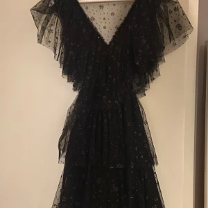 Svart klänning med volang från Ida Sjöstedt - Elegant svart klänning från Ida Sjöstedt med v-ringning och volangdetaljer. Klänningen har ett glittrigt blommönster och är tillverkad i ett transparent material som ger en lyxig känsla. Perfekt för speciella tillfällen. Original pris 5595 och säljs inte längre, använd en gång men säljer då den inte är min stil 