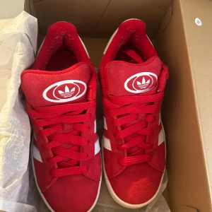 Röda sneakers från Adidas campus  - Säljer ett par snygga röda Adidas sneakers i mycket bra skick. De har en klassisk design med vita detaljer och snörning. Perfekta för både vardag och fest! Storleken är normal och de är gjorda i syntetmaterial. Nästan nya använd två gånger.