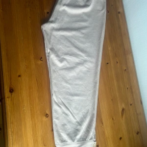 Beige byxor från Shein - Säljer ett par bekväma beige byxor från Shein i storlek 2XL. Byxorna har en mjuk och stretchig känsla, perfekt för en avslappnad stil. De har knappar vid bensluten som en snygg detalj. Passar perfekt för en chill dag hemma eller en casual utflykt.