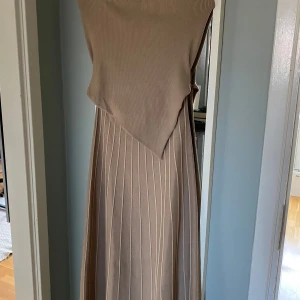 Beige ribbad klänning från ADOORE.  - Höst-nyhet från Adoore! Lausanne bandeau dress Snygg beige ribbad klänning i ett mjukt material. Den har en asymmetrisk design med en unik omlott-look framtill. Perfekt för både vardag och festliga tillfällen. Klänningen är ärmlös och har en lång längd som ger en elegant siluett.                